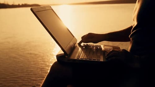A man is sitting and working on the laptop near the lake in the background of the sunset