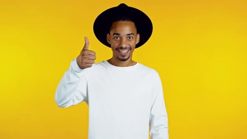 Hübscher afrikanischer Mann mit Hut zeigt Daumen hoch, wie eine Geste. Glücklicher Kerl auf gelbem Hintergrund.
Gewinner