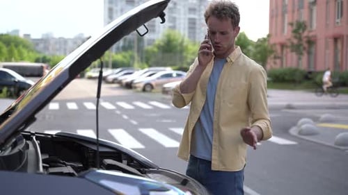 Man stands near a car with an open hood and talks on the phone.