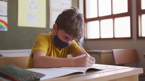 Boy wearing face mask writing while sitting on his desk at school