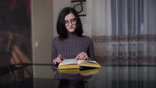 Serious young girl reads a book while sitting in the kitchen in the evening