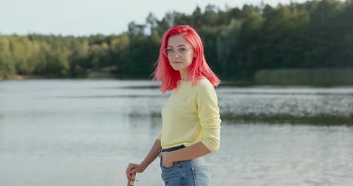 Cheerful Girl with Pink Hair Blown By the Wind Wearing Glasses Stands Near the Lake Hand Resting on