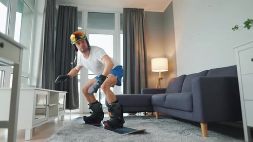 Vídeo divertido. Homem de bermuda e camiseta retrata snowboard em um tapete em um quarto aconchegante. Esperando por