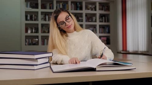 Ein Studentenmädchen mit Brille sitzt in einer Bibliothek und schreibt in ein Notizbuch