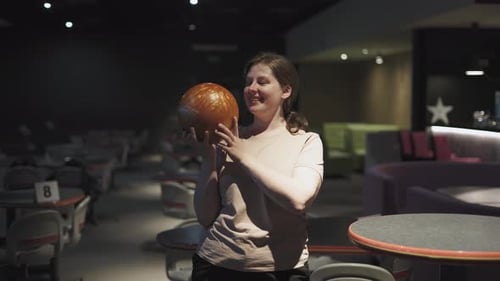 Charming Young Woman Stands with Ball in Dark Hall of the Bowling Alley