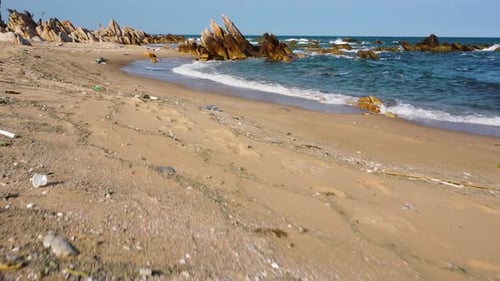 Powerful waves hit the shore polluted with plastic trash. Rocks rise above the sandy beach