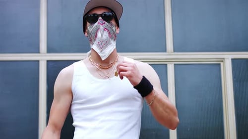 Adult man with golden necklaces around his neck and scarf over face makes W with his hand.