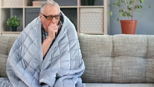 Senior Man Coughing on Couch Wrapped in Blanket