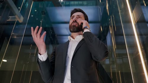 Charismatic Successful Businessman Riding Glass Elevator Talking on Mobile Phone