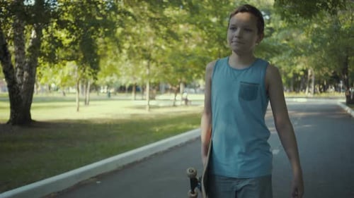 Skateboarder Walks in a Park with a Skateboard in Hands.