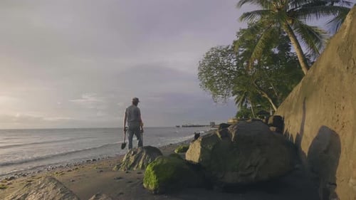 Man Is Scanning Sand Beach By Metal Detector and Walking with His Dog at Sunset Near the Ocean.