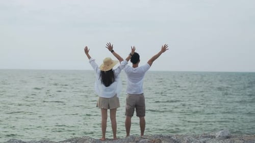 Couple Raising Arms Facing Ocean on Rocky Breakwater