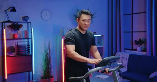 Chico asiático deportivo con ropa de entrenamiento que marca la velocidad en el panel de control de la cinta de correr durante la noche