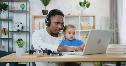 Black-Skinned Guy in Headset Holding His Small Son on Knees while Working on Computer at Home