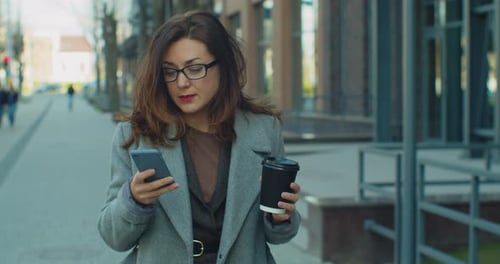 Successful Business Woman with Glasse Walking with Coffee and Uses Smartphone in Business District