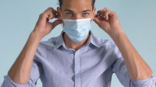 Caucasian Man Demonstrating Proper Wearing Of Surgical Mask In Plain Blue Background - medium close