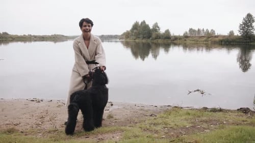 An Elegant Young Brunette in a White Coat Stands By the River and Holds a Large Black Poodle on a