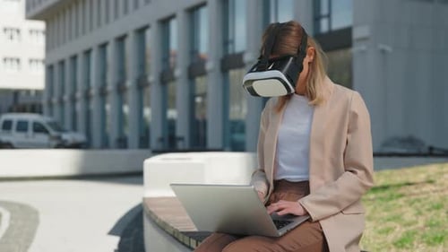 Woman in VR Headset Sitting with Laptop Near Office Building