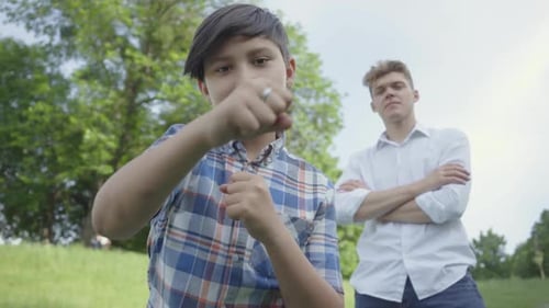 The Boy Punching Air Looking in the Camera in the Summer Park. His Father of Older Brother Standing