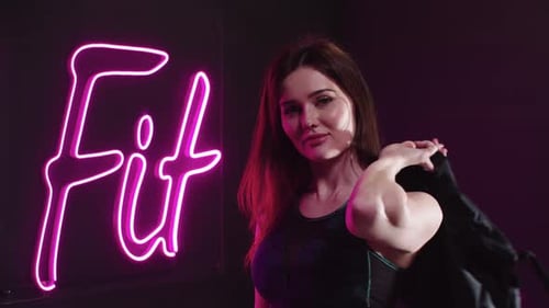 Young Pretty Woman in Sportive Top Walks to the Neon Sign Saying ''Fit'' and Looks in the Camera