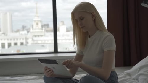 A Young Blonde Woman Works Behind a Laptop Near a Large Window