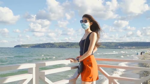Mulher em forma com óculos de sol e máscara facial olhando por cima de uma cerca no mar. Medidas de proteção durante