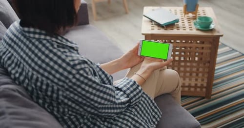 Girl Looking at Green Mock-up Screen of Smartphone Sitting on Sofa at Home