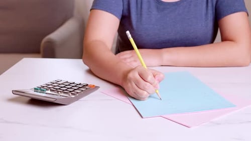 Woman is using a calculator to calculate and write down her monthly accounting expenses at home.