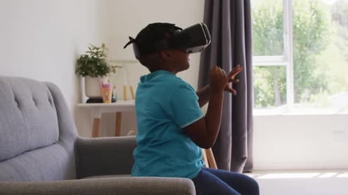 African american boy gesturing while using vr headset sitting on the couch at home