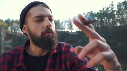 Portrait d un barbu assis dans la forêt près de la rivière et fumant un cigare brun.