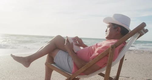 Latin senior hispanic man relaxing on sunbed on beach at sunset, reading book