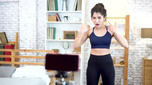 Mujer asiática en ropa deportiva hablando con la cámara y explicando tutoriales de acondicionamiento físico en línea en la sala de estar