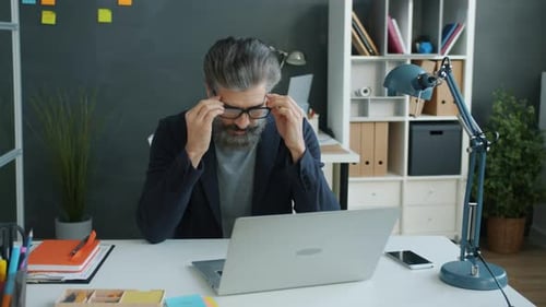 Slow Motion of Serious Mature Man Putting on Glasses Then Working with Laptop in Office