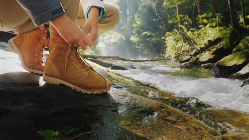 Female Hands Lace Up the Female on a Trekking Boot in the Forest. In the Background, a Mountain