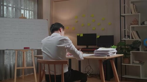 Back View Of Asian Man Writing Something On Paper While Working With Documents At The Office