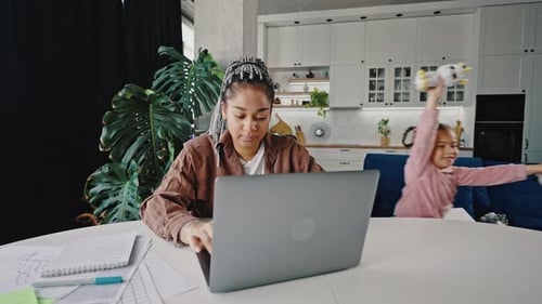 Young African American Woman Working on Laptop Her Active Little Daughter Running Around and
