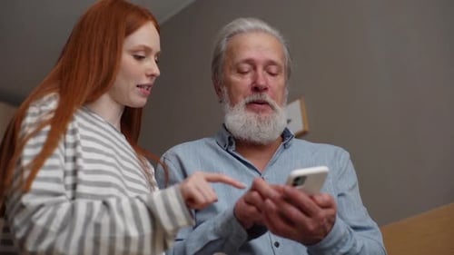 Lowangle View of Bearded Senior Grandfather Learning to Using Mobile Phone Under Guidance of Pretty