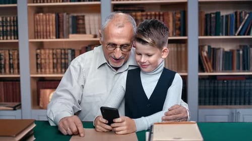 Grandchild Teaching Grandfather Use Smartphone Chatting Surfing Internet at Table in Home Library