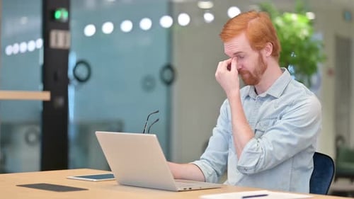 Stressed Young Redhead Man with Laptop Having Headache in Office