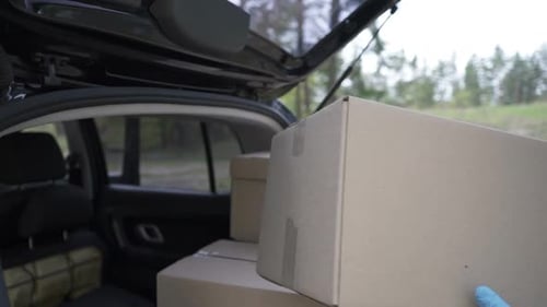 A Volunteer in a Protective Suit Loads Boxes Into the Trunk of a Car