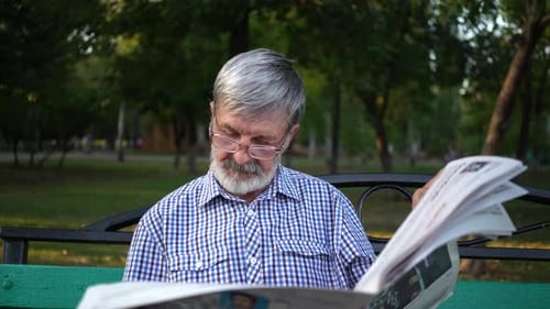 Senior in Plaid Shirt Sits on a Bench in the Park and Reads a Newspaper