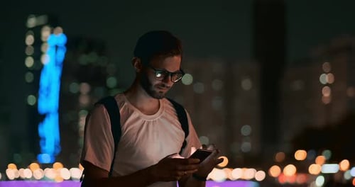 A Man in Glasses Against the Background of the Night City Writes a Message Using a Smartphone