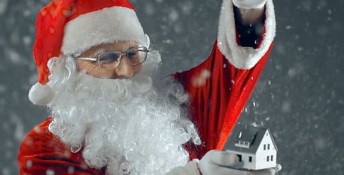 Santa Claus Holding a Tiny House Under Falling Snow
