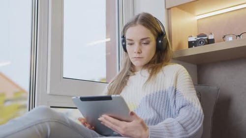 Jeune femme assise près de la fenêtre à la maison dans un casque, travaillant sur une tablette. Étude à distance, travail.