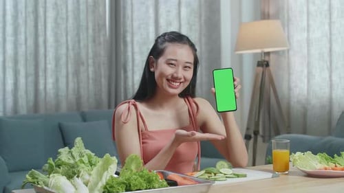 Asian Woman With Healthy Food Holding And Pointing Green Screen Smartphone While Sitting At Home