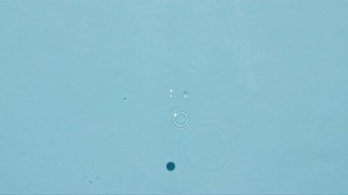 Top View Slow Motion of Drop Falls Into Water and Diverging Circles of Water on Blue Background