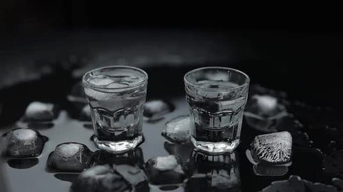 Add Ice Cubes To Shots of Vodka in Glass Against Black Background. Alcohol Drink