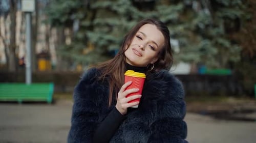 Portrait of a model outdoors. Elegant woman in fur coat holding plastic glass and looking on camera
