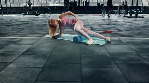 A flexible girl athlete rolls her muscles with a roller on a karemat in the gym.