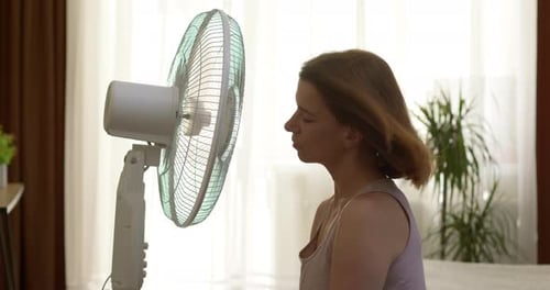 A woman sits near a rotating fan in a stream of cool air in a hot room in summer.